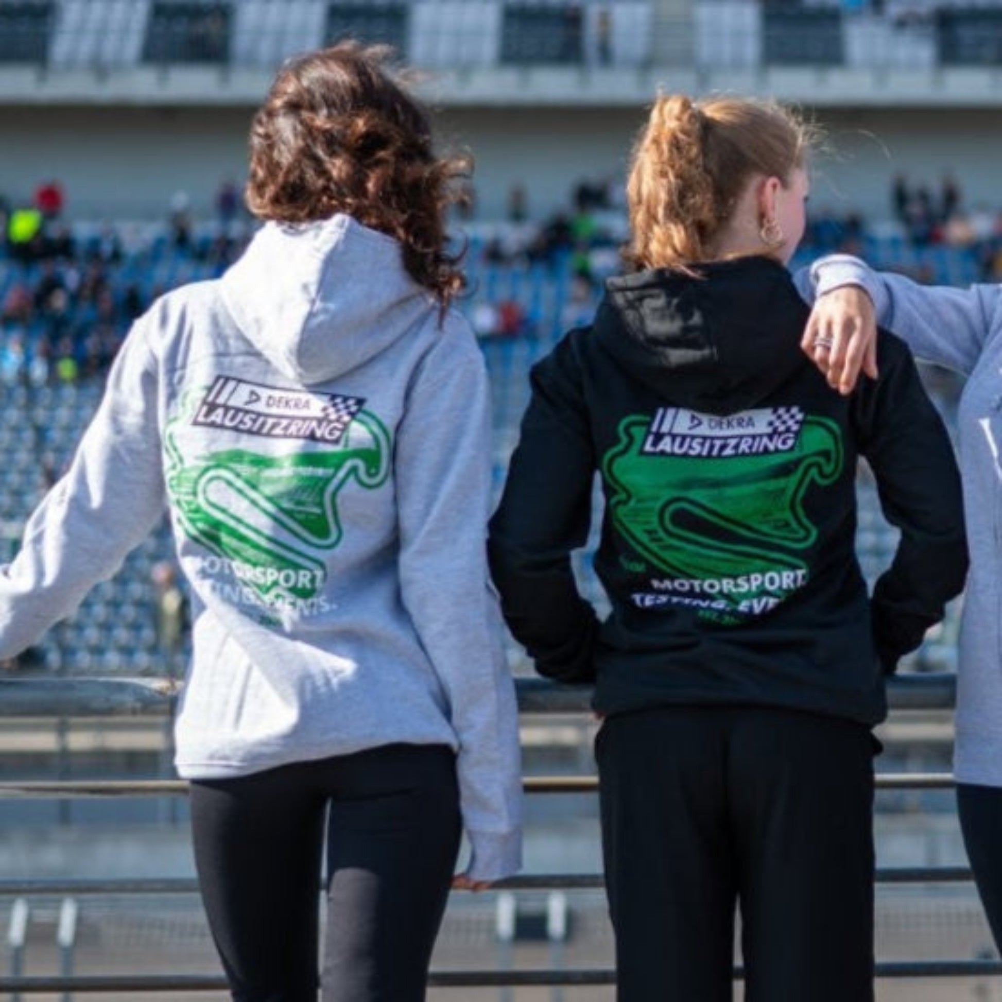 Rückansicht mehrerer Personen im Zip Hoodie Motorsport, Testing & Events mit grünem DEKRA Lausitzring Logo, sportliche Teamwear für Fahrer und Fans
